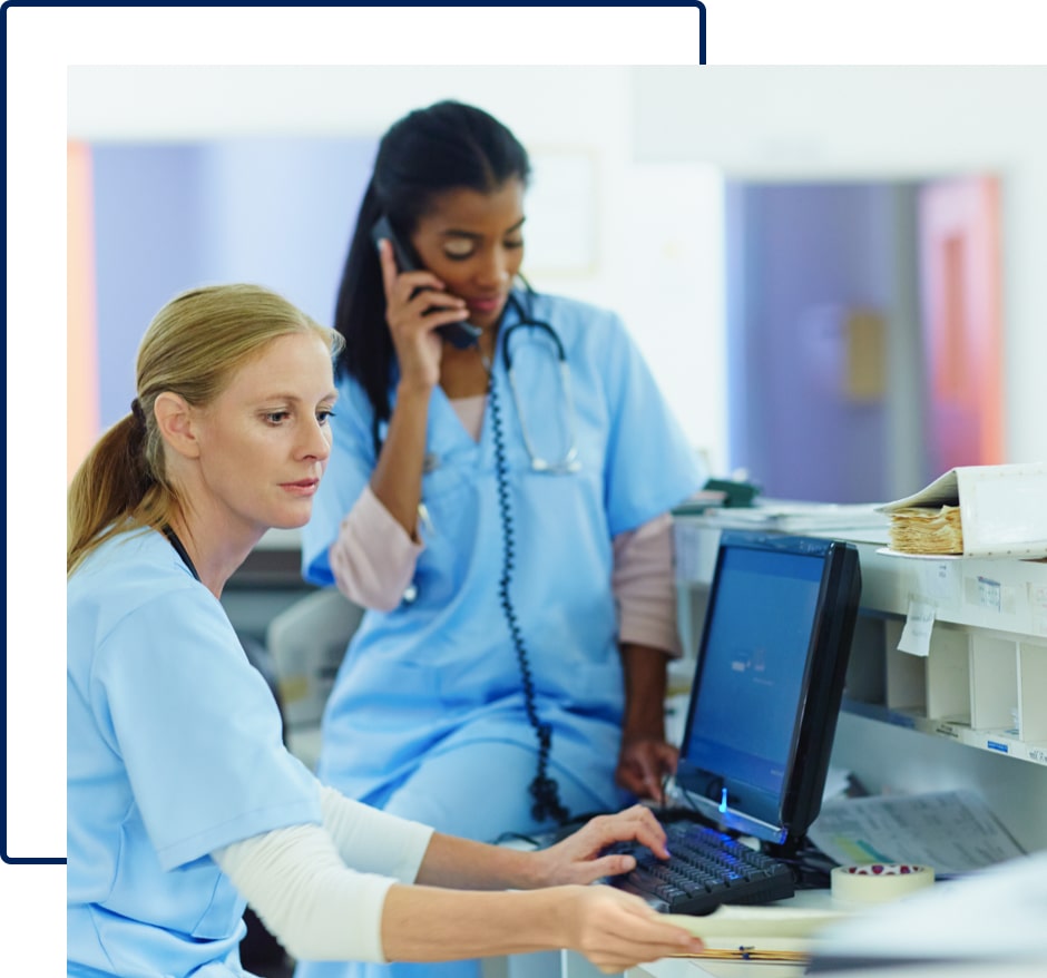 One female doctor is sitting and working on computer and the other one is talking over a phone.